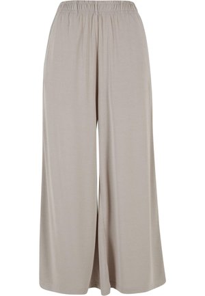 Frau trägt beige hoch taillierte, weit geschnittene Hose, schwarzes kurzes Tanktop und weiße Sneakers, steht vor einfachem grauem Hintergrund.