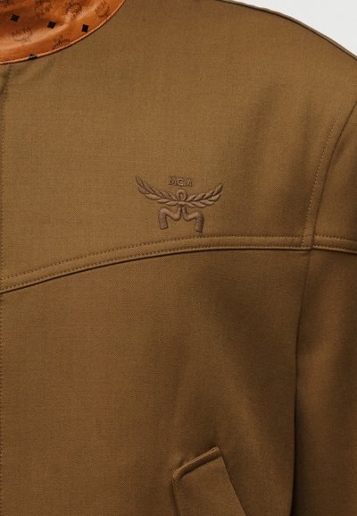 Καφέ ζακέτα με κεντημένο λογότυπο MCM που φέρει έμβλημα με φτερά και μικρό δερμάτινο κολάρο με ανάγλυφα σχέδια.