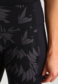 Lähikuva mustista legginsseistä, joissa on tummat geometriset kuviot ja pieni sivutasku, joka on henkilön lantiolla ja reidessä.