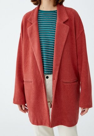 Femme portant un blazer en velours côtelé couleur rouille surdimensionné, avec une chemise rayée bleu sarcelle et blanc, et un pantalon beige clair, debout devant un fond blanc.