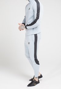 Světle modrý sportovní komplet s černými bočními pruhy, vyrobený z texturované tkaniny; obsahuje límec na půl zip a přiléhavé kalhoty. Zahrnuté černé obuv.