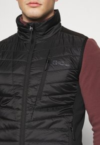 Sort polstret vest med høj krave, der har en lynlåslomme på brystet. Blødt stof med branding på venstre side.