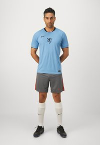 Maglia Nike azzurro chiaro con logo di leone nero, pantaloni corti grigi con accenti rossi, calze bianche e scarpe sportive nere.