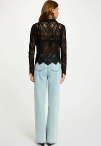 Top lungo in pizzo nero con maniche lunghe e orlo scallopato, caratterizzato da intricati motivi a diamante. Indossato con jeans a vita alta, larghi e di colore blu chiaro.