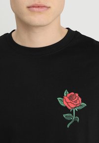 Muž v černé košili s červenou růží a zelenými listy potisknutými na levé hrudi, na jednobarevném světlém pozadí.