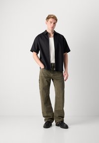 Short-sleeve black shirt with zip closure, worn over a white ribbed tank top. Olive green pants with side pockets and black footwear.