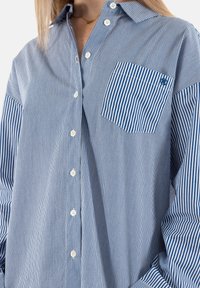 Camisa de botonera a rayas azul y blanca con un bolsillo en el pecho, que cuenta con un cuello clásico y una tela texturada.