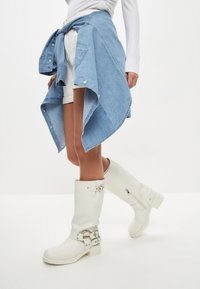 Białe wysokie kozaki za kolano z zapięciami na klamry i detalem zamka, w połączeniu z jasnoniebieską denimu koszulą w stylu wrap i białymi szortami.