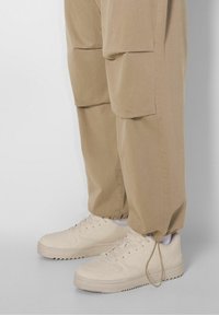 Beige lenkkarit matalalla varrella, kumipohjalla, rei'itetyillä yksityiskohdilla ja tasaisilla nauhoilla, yhdistettynä väljään ruskeaan housuun, jossa on sivutaskut.