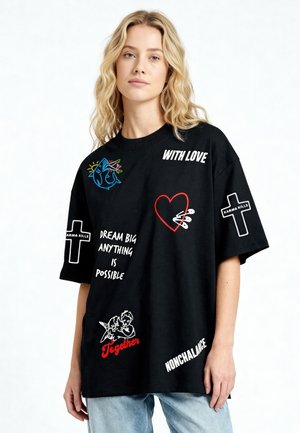 Donna bionda che indossa una T-shirt nera oversize con frasi, croci, un cuore con spille, angeli e un disegno di uno squalo.