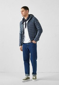 Navyblauwe pufferjack met capuchon, lichtgewicht stof, geopend. Witte t-shirt, blauwe spijkerbroek, groene schoenen met witte veters, grijze sjaal.