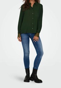 Mørkegrøn skjorte med lange ærmer og blondeaccenter; parret med blå skinny jeans og sorte ankelstøvler.