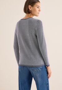 Grauer Strickpullover mit strukturiertem Muster, Rundhalsausschnitt, langen Ärmeln und geripptem Saum. Kombiniert mit blauen Jeans.
