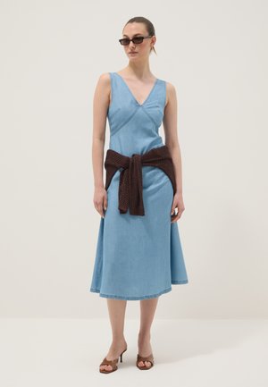 Frau trägt ein hellblaues ärmelloses Midikleid mit einem dunkelbraunen Strickpullover, der um die Taille gebunden ist, braune Sandaletten mit Absatz und schwarze Sonnenbrille.