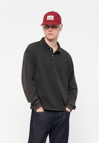 Svart langermet polo skjorte med krage og tre knapper, kombinert med mørkeblå jeans og en burgunder caps med en logo-patch.