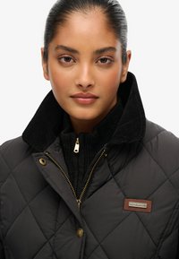 Kvinde med mørkt hår i en høj knold iført en sort quiltet jakke med en høj corduroy krave og en guld lynlås.