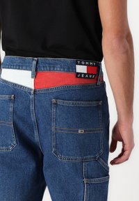 Jeans en denim avec une finition bleu foncé. Les caractéristiques incluent une taille bicolore en rouge, blanc et bleu, ainsi que deux poches arrière.