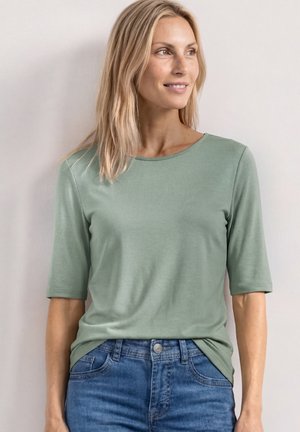 Blonde Frau trägt ein grünes kurzärmeliges Shirt und blaue Jeans, schaut seitlich mit einem leichten Lächeln, steht vor einem einfarbigen Hintergrund.