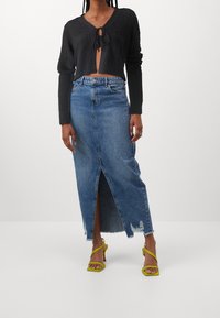 Černý krátký svetr s předním zavazováním a texturovaným pletením, spárovaný s bledě modrou denimovou maxi sukni s předním rozparkem a třepícími se okraji. Jasně žluté sandály na tenkých ramínkách.