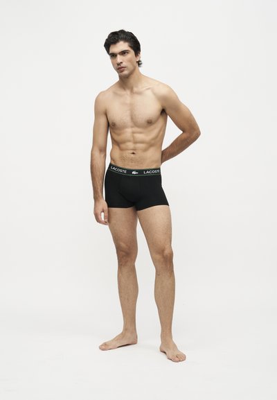Jeune homme debout, pieds nus, torse nu, portant un boxer noir Lacoste avec une ceinture logo sur fond blanc uni.