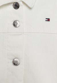 Camicia in denim bianco con bottoni a pressione argento, cuciture visibili e un piccolo logo ricamato in blu, rosso e bianco.