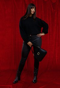 Suéter negro oversized, pantalones de cuero negros, botines negros con tacón y un bolso negro con detalle de hebilla dorada contra un fondo rojo.