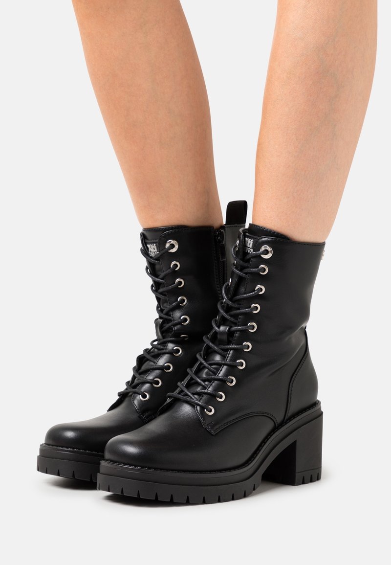Bottines montantes en cuir noir à lacets avec talons épais portées sur des jambes nues sur un fond neutre.