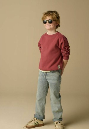 Enfant aux cheveux blonds ébouriffés portant des lunettes de soleil noires, un pull rouge, un jean bleu clair et des baskets beige, debout les mains derrière le dos.