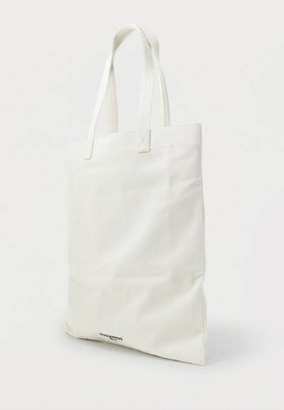 Λευκή τσάντα tote καμβά με διπλούς ιμάντες και μικρό μαύρο λογότυπο μάρκας κοντά στην κάτω άκρη, στέκεται όρθια σε απλό φόντο.