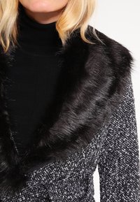 Collar de piel sintética negra en un abrigo de textura gris y negro con un patrón. El primer plano muestra la suavidad del collar y el detalle tejido del abrigo.