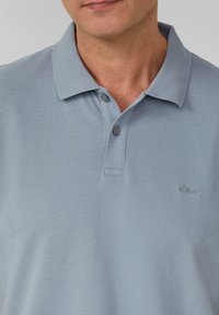 Hellblaues Poloshirt aus strukturiertem Stoff, kurzer Kragen und drei Knöpfen, mit einem dezenten Logo auf der linken Brust.