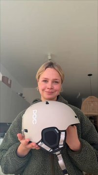 Personne souriante tenant un casque beige POC à l'intérieur, vêtue d'une veste polaire verte, avec un intérieur de maison moderne en arrière-plan.