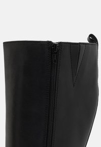 Botte en cuir noir avec une fermeture éclair latérale, une texture lisse et un design structuré. Détail de couture remarquable le long du bord supérieur.