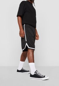 Shorts de sport noirs avec des rayures blanches sur les côtés, associés à un t-shirt noir et des baskets noires. Chaussettes blanches visibles au-dessus des chaussures.