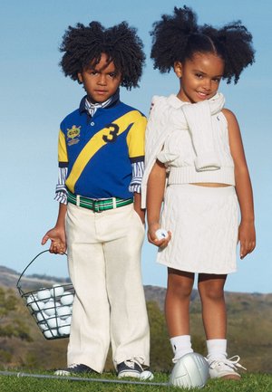 Δύο παιδιά στέκονται στο γρασίδι με μπάλες γκολφ· το αγόρι κρατάει ένα καλάθι με μπάλες, το κορίτσι κρατάει μια μπάλα, ένα γκολφ κλαμπ βρίσκεται στα πόδια τους κάτω από καθαρό ουρανό.