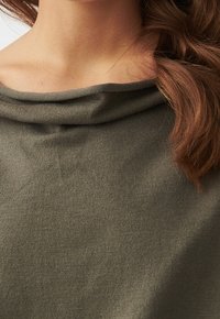 Femme portant un pull en tricot vert olive avec un col large et décontracté, et des cheveux bruns ondulés tombant sur une épaule.