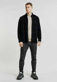 Camicia di flanella a quadri neri sopra una felpa beige chiaro, abbinata a pantaloni cargo grigi aderenti e sneakers nere con dettagli bianchi.
