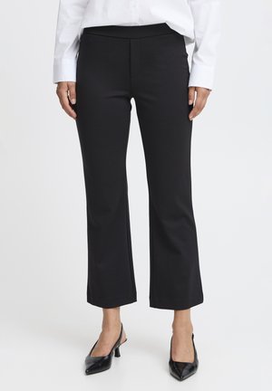 Femeie purtând pantaloni evazați negri croppați, cămașă albă cu mâneci lungi și pantofi slingback negri cu vârf ascuțit, stând în fața unui fundal alb.