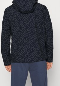 Námořnicky modrá bunda s kapucí s geometrickým vzorem v světlejší šedé. Texturovaná látka, dlouhé rukávy a těsné zakončení u lemu.