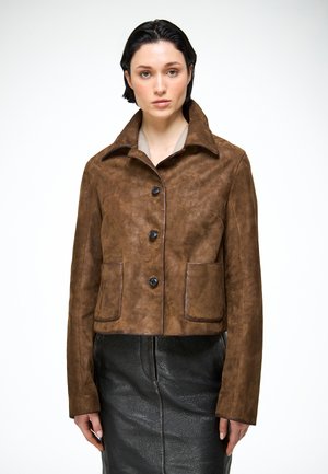 Mujer con cabello corto y oscuro, vestida con una chaqueta de ante marrón con botones y una falda de cuero negra, de pie contra un fondo blanco.