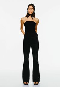 Schwarzes, trägerloses Peplum-Oberteil, kombiniert mit schwarzen, weit ausgestellten Jeans. Das Model trägt ein schwarz-weiß gemustertes Kopftuch und schwarze Flip-Flops.