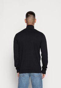 Pull noir à manches longues avec un col montant, fabriqué dans un tissu lisse, avec des poignets et un ourlet côtelés, porté avec un jean bleu clair.