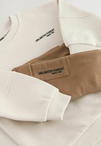 Sweatshirt beige avec des poignets côtelés, comportant le texte noir "UNLIMITED FUNDAY SINCE 1982". Manche marron posée sur le tissu beige.