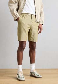 Σορτς ανοιχτό ελιά χρώματος, κατασκευασμένα από υφασμάτινη ύφανση με υφή, διαθέτουν ελαστική μέση με κορδόνι. Συνδυάζονται με μπεζ bomber jacket και αθλητικά παπούτσια.