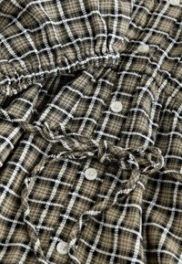 Tessuto a quadri marrone e nero con accenti bianchi, caratterizzato da una vita arricciata, bottoni e una consistenza morbida, che mette in mostra un design stropicciato.