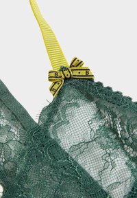 Zielony koronkowy biustonosz typu bralette z falistym dołem, regulowanym żółtym paskiem oraz ozdobną kokardką na środku z czarnym napisem.