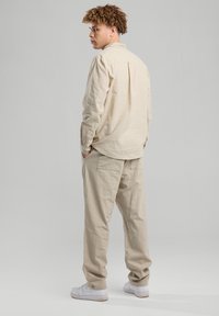 Beige liinapaita, jossa on kaulus, pitkät hihat ja takana yoke-yksityiskohta, yhdistettynä löysiin beigeihin housuihin ja valkoisiin lenkkitossuihin.