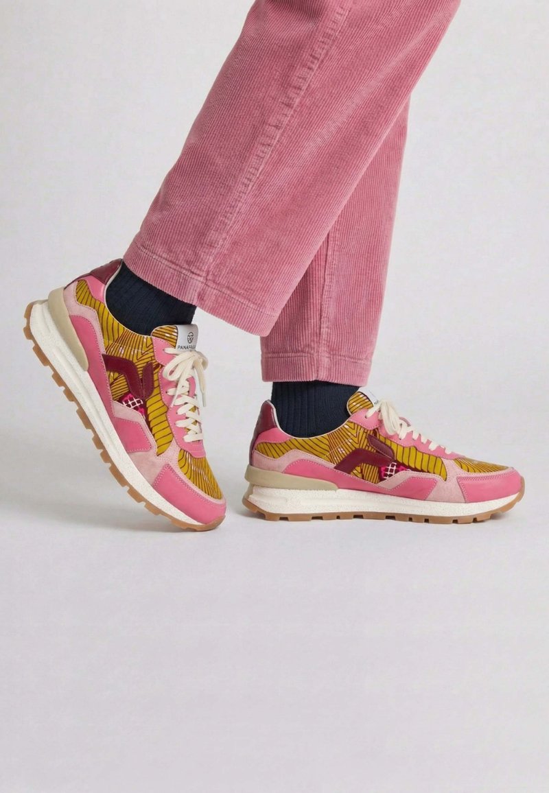 Personne portant un pantalon en velours côtelé rose et des baskets multicolores rose et jaune avec des chaussettes noires, sur un fond gris uni.