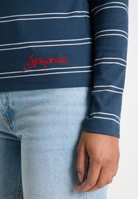 Langärmliges Oberteil mit Marine-Streifen, weißen Streifen und rotem besticktem Text auf der Vorderseite, kombiniert mit hellblauen Denim-Jeans.