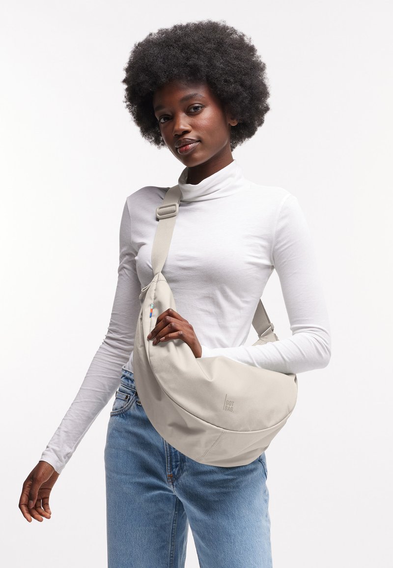 Mujer con peinado afro natural que lleva un suéter de cuello alto blanco y vaqueros azules, sosteniendo un bolso cruzado beige sobre su torso.